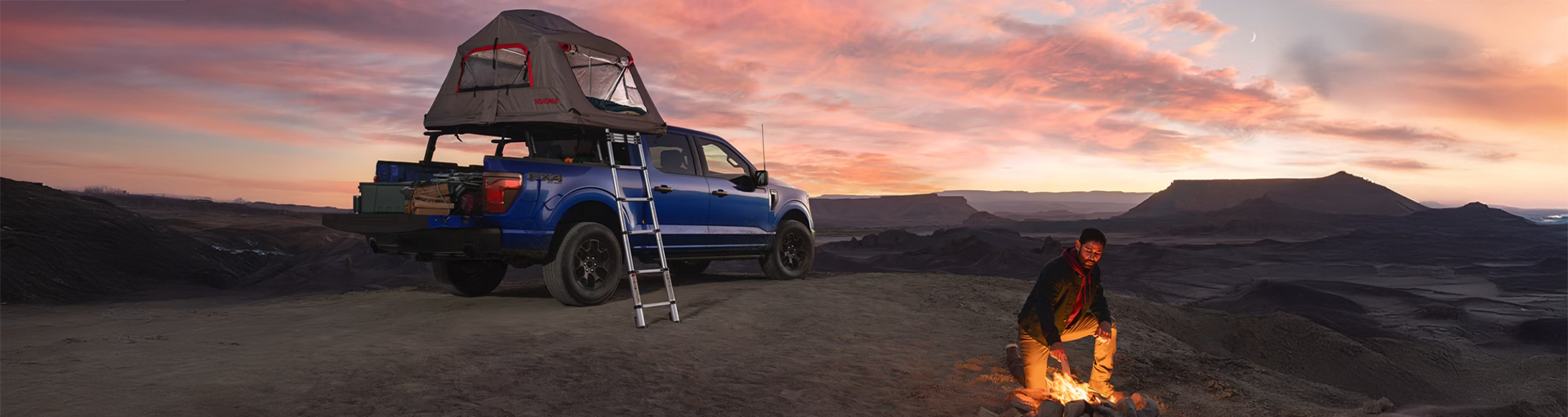 A blue Ford F-150 parked off road at a camp site with camping accessories attached and a man sitting by a camp fire.