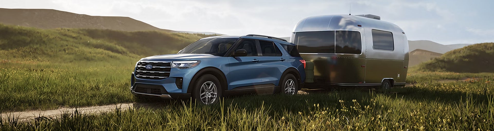 A blue Ford Explorer towing a silver travel trailer along a grassy dirt path.